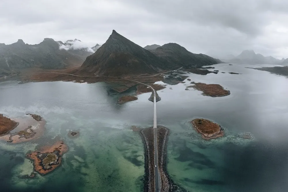 Aerial view of a road on a narrow causeway over flat, turquoise water with small islands, a pointed mountain in the mist behind