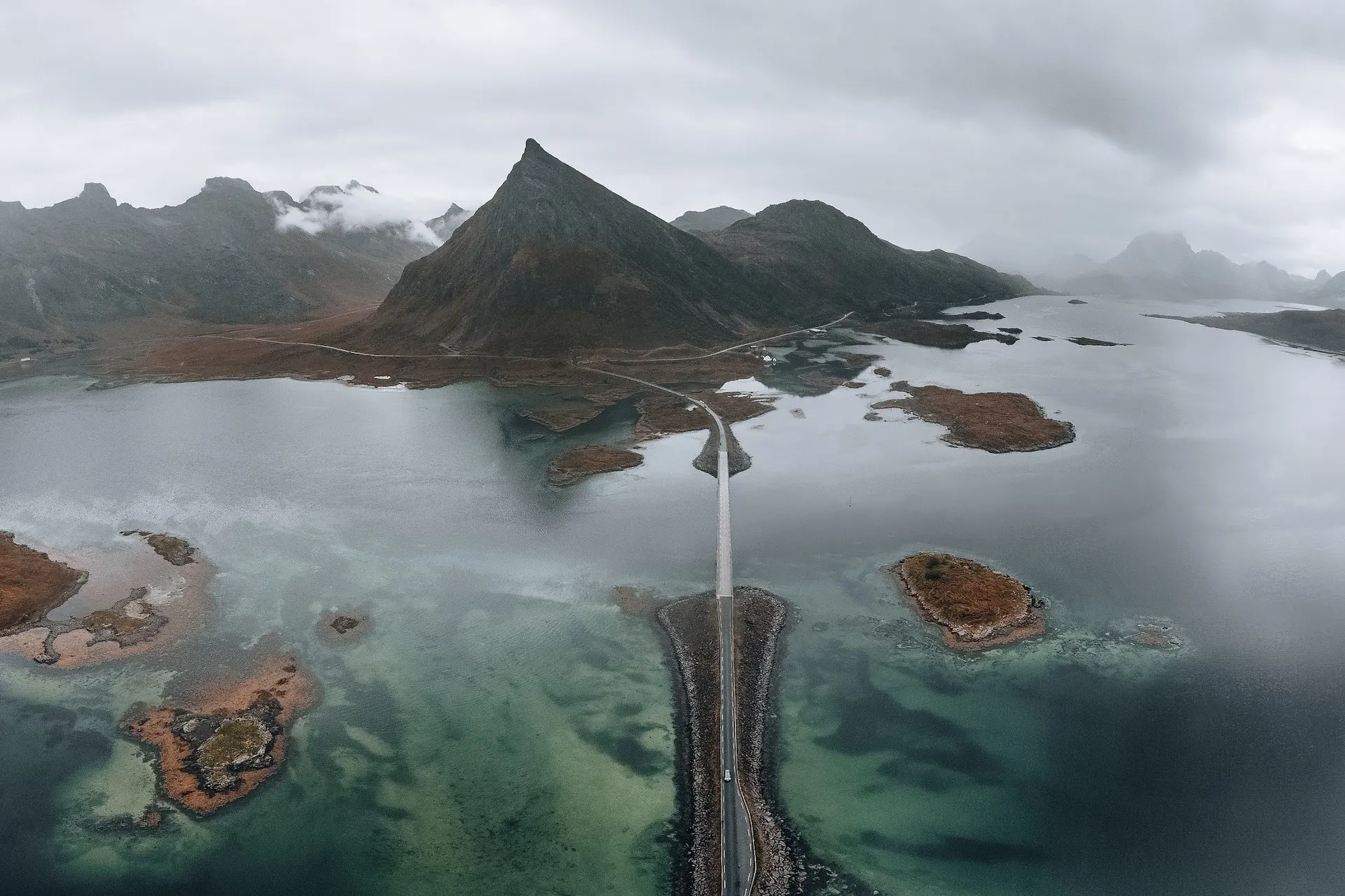 Luftaufnahme einer Strasse auf einem schmalen Damm über flaches, türkises Wasser mit kleinen Inseln, dahinter ein spitzer Berg im Nebel