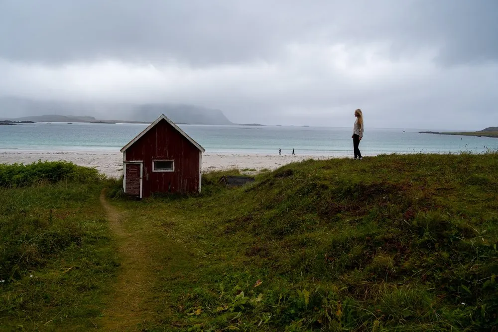 The red wooden cabin at Rambergstranda, a person stands on the grassy hill beside it looking out to sea