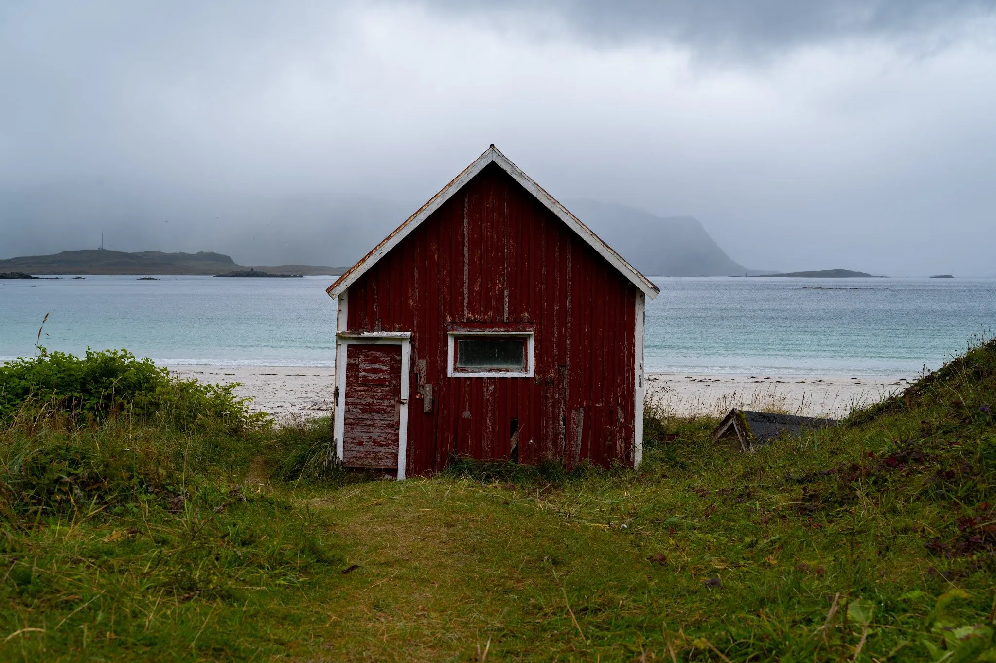 The weathered red wooden cabin stands between grass and sandy beach at Rambergstranda, the sea behind it under heavy clouds
