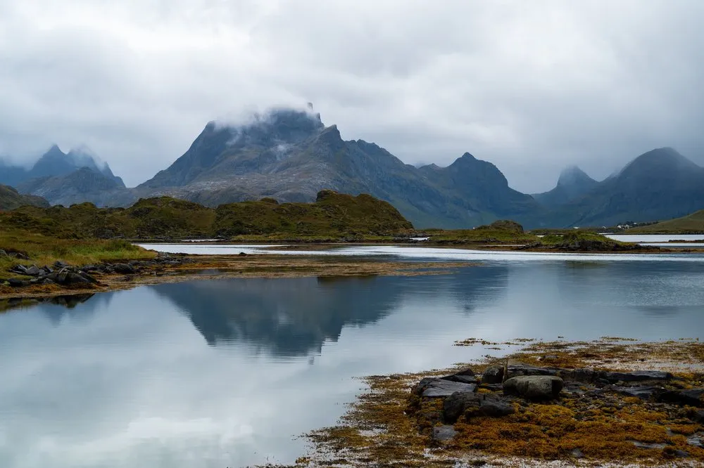 A mountain range with cloud-covered peaks is reflected in a still bay, stones and seaweed in the foreground