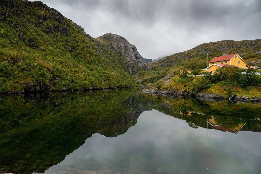 A yellow house with a red roof stands on the slope above mirror-smooth water, an overgrown rock wall to the left