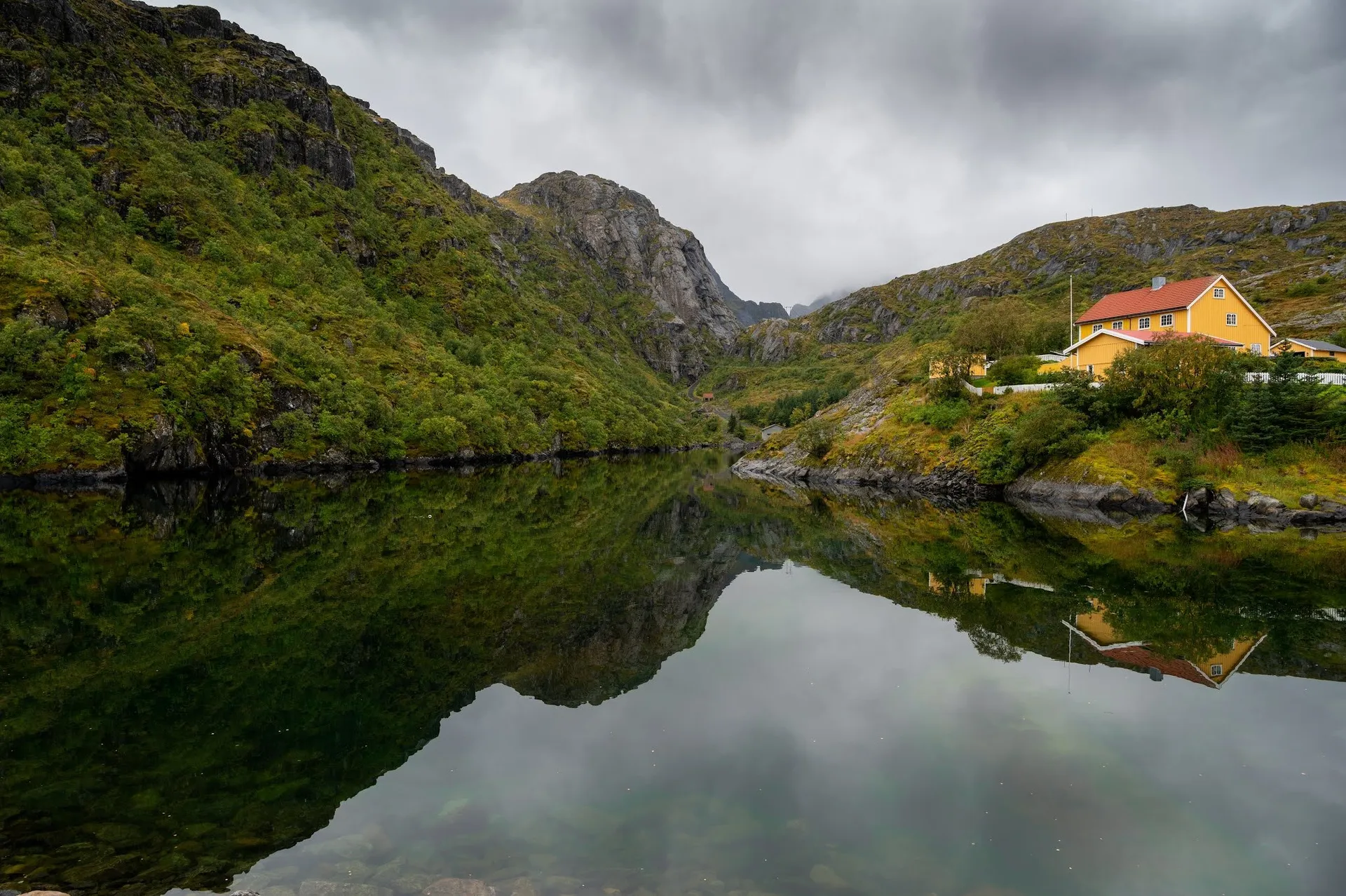 Ein gelbes Haus mit rotem Dach steht am Hang über spiegelglattem Wasser, links eine bewachsene Felswand
