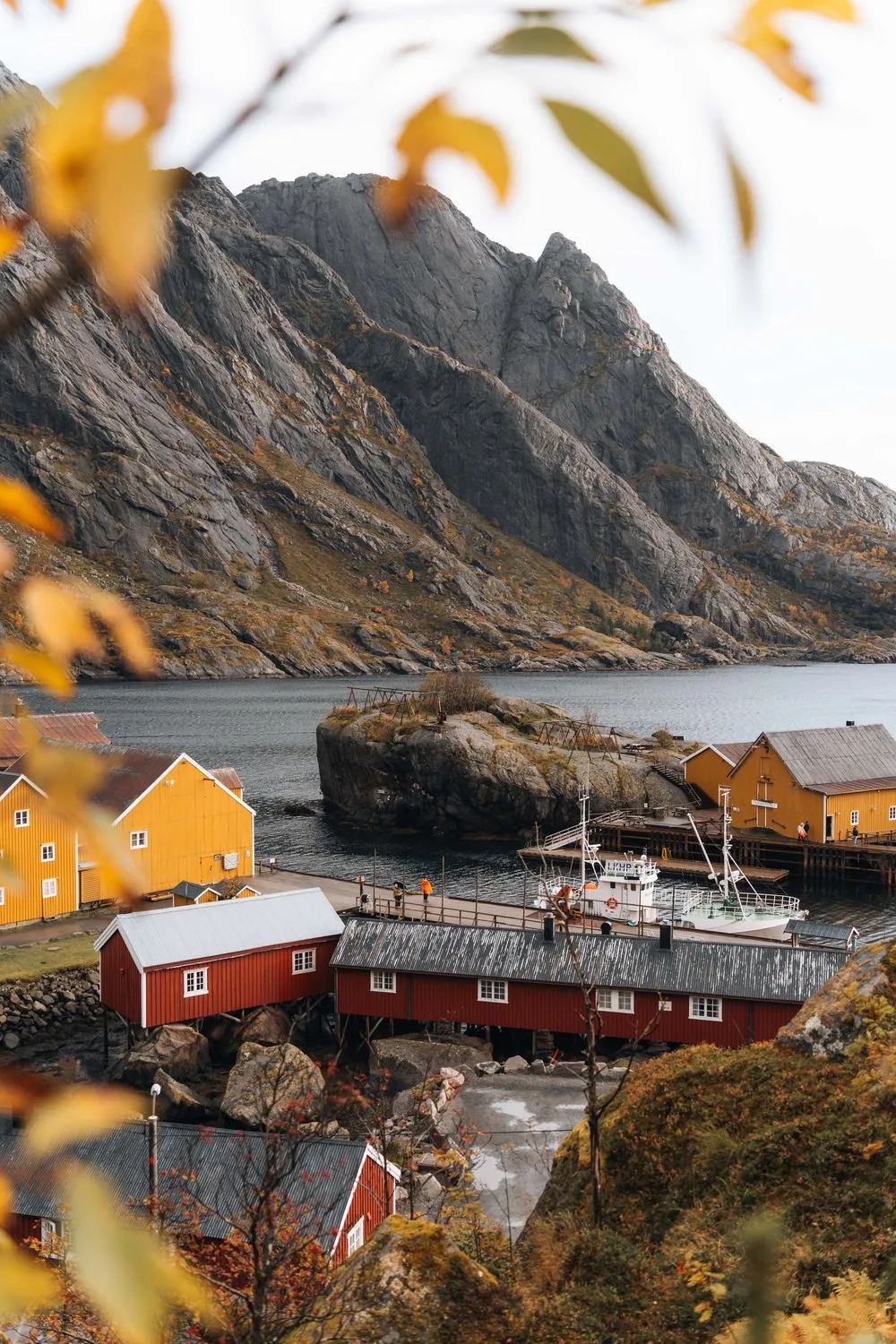 Gelbe und rote Häuser von Nusfjord von oben, im Vordergrund herbstliches Laub