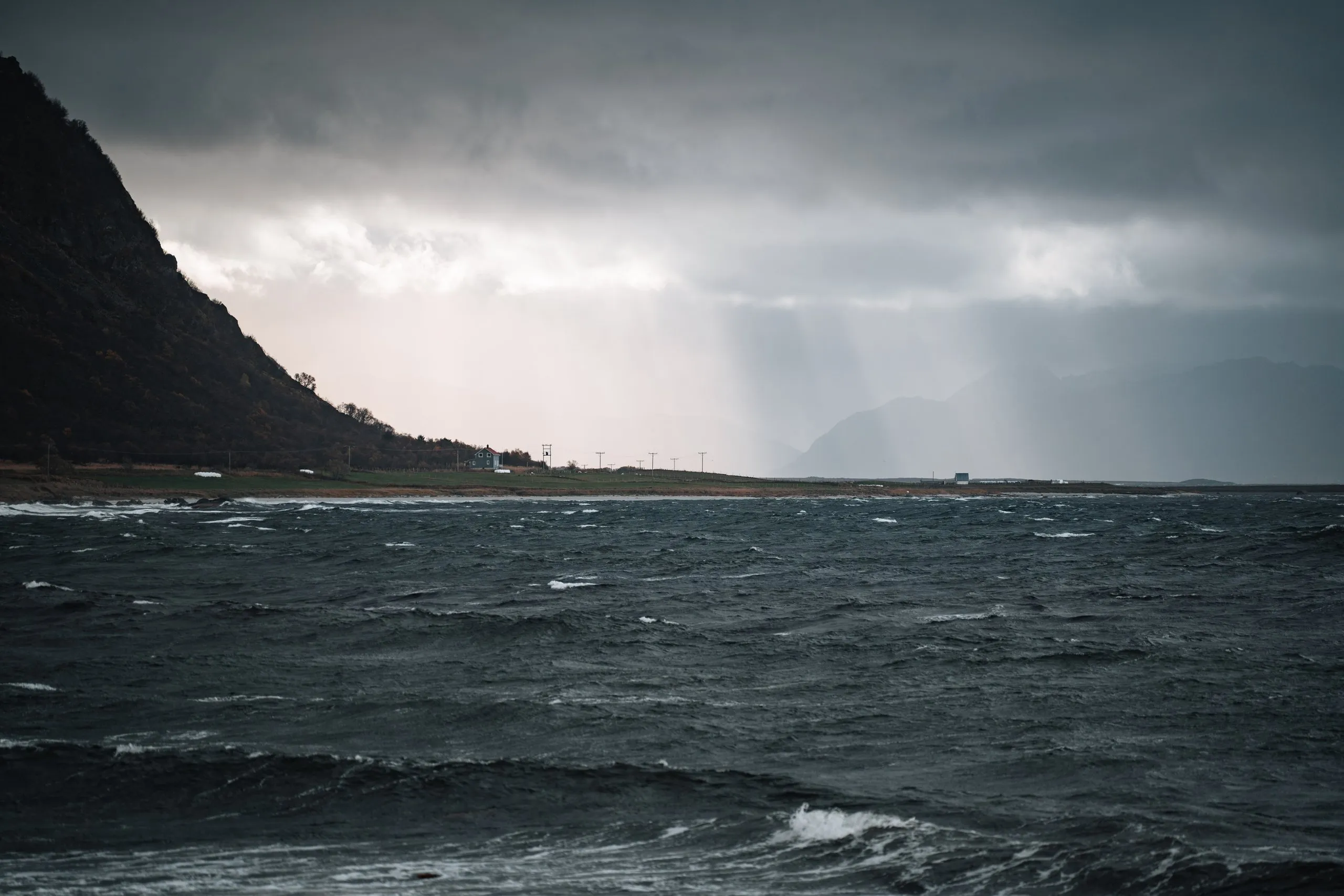 Rain showers hang like bright veils over the rough sea at Nøss, a dark mountainside on the left, a single house on the shore