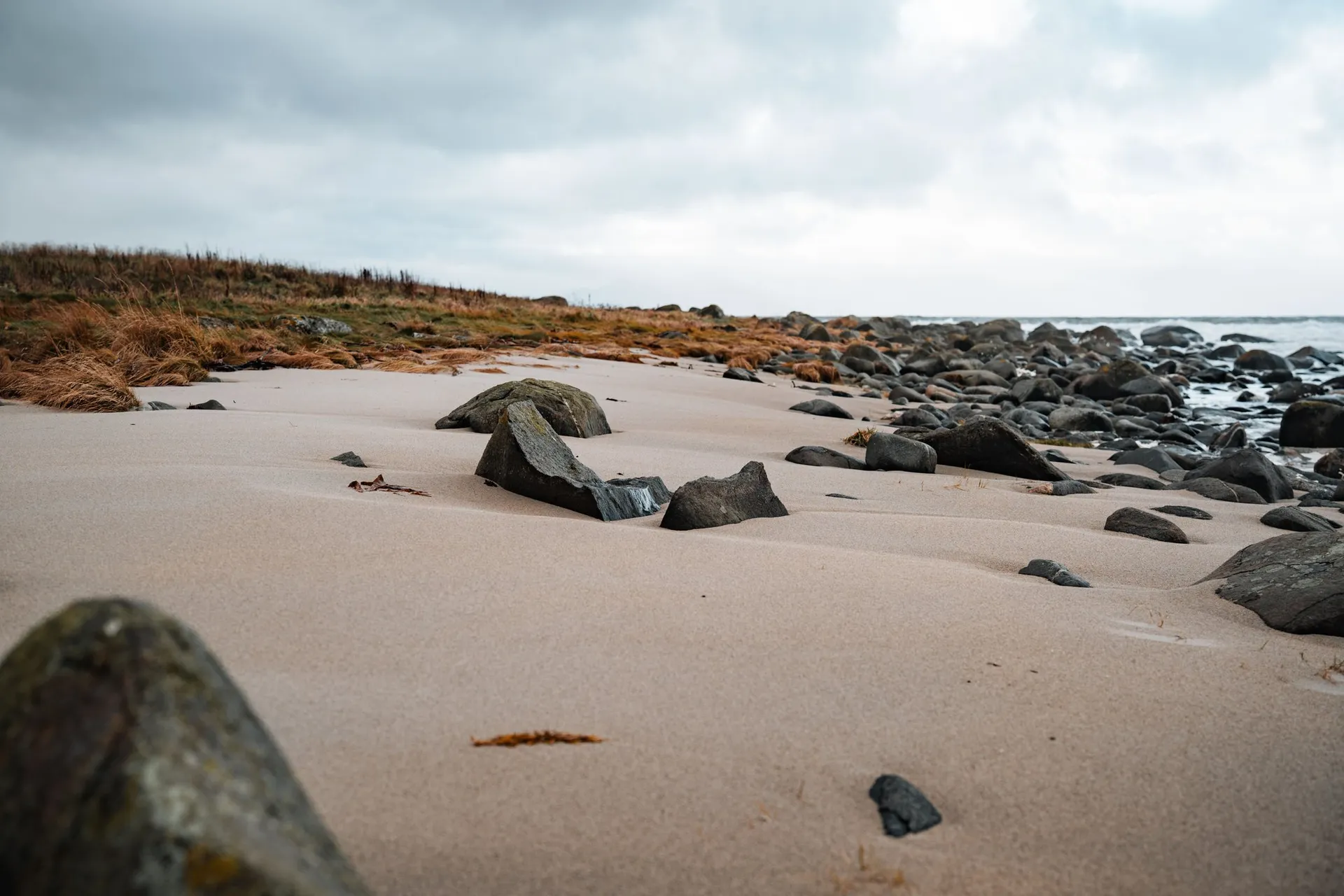 Dark round stones in the light sand on the beach at Nøss, behind them dry brown grass under a grey sky