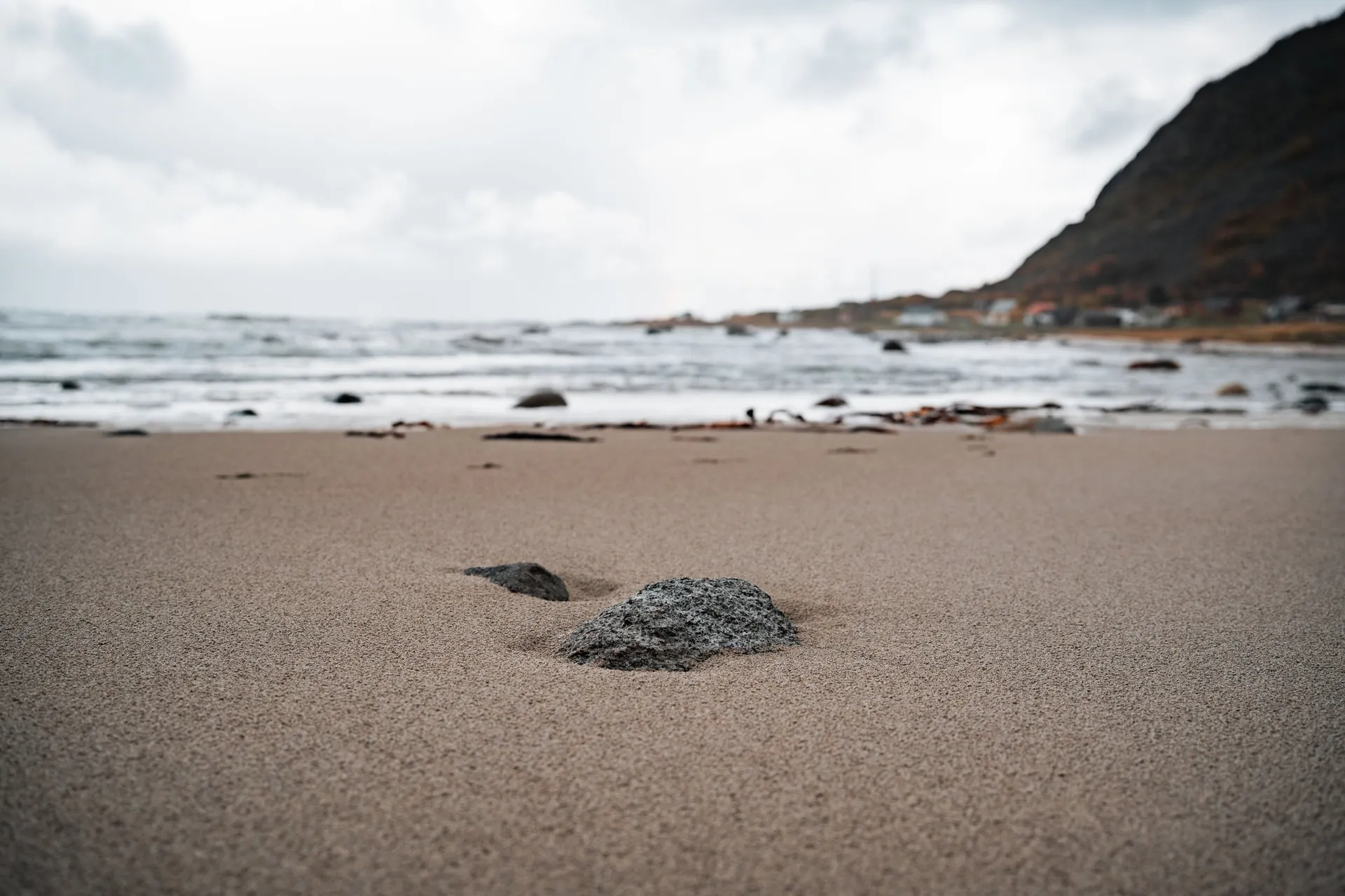 A grey stone in the fine sand on the beach at Nøss, behind it foaming waves and a dark mountain with houses at its foot
