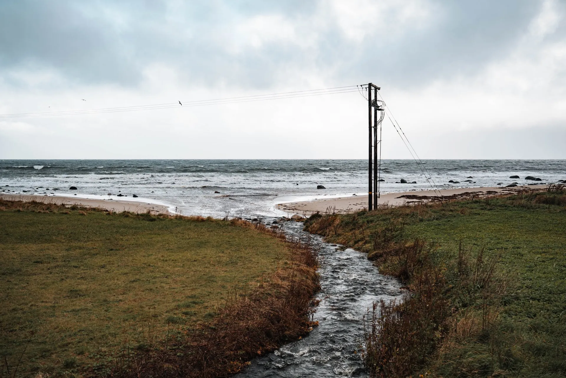 A stream runs between wet meadows to the beach at Nøss, next to it a wooden power pole, behind it the rough sea