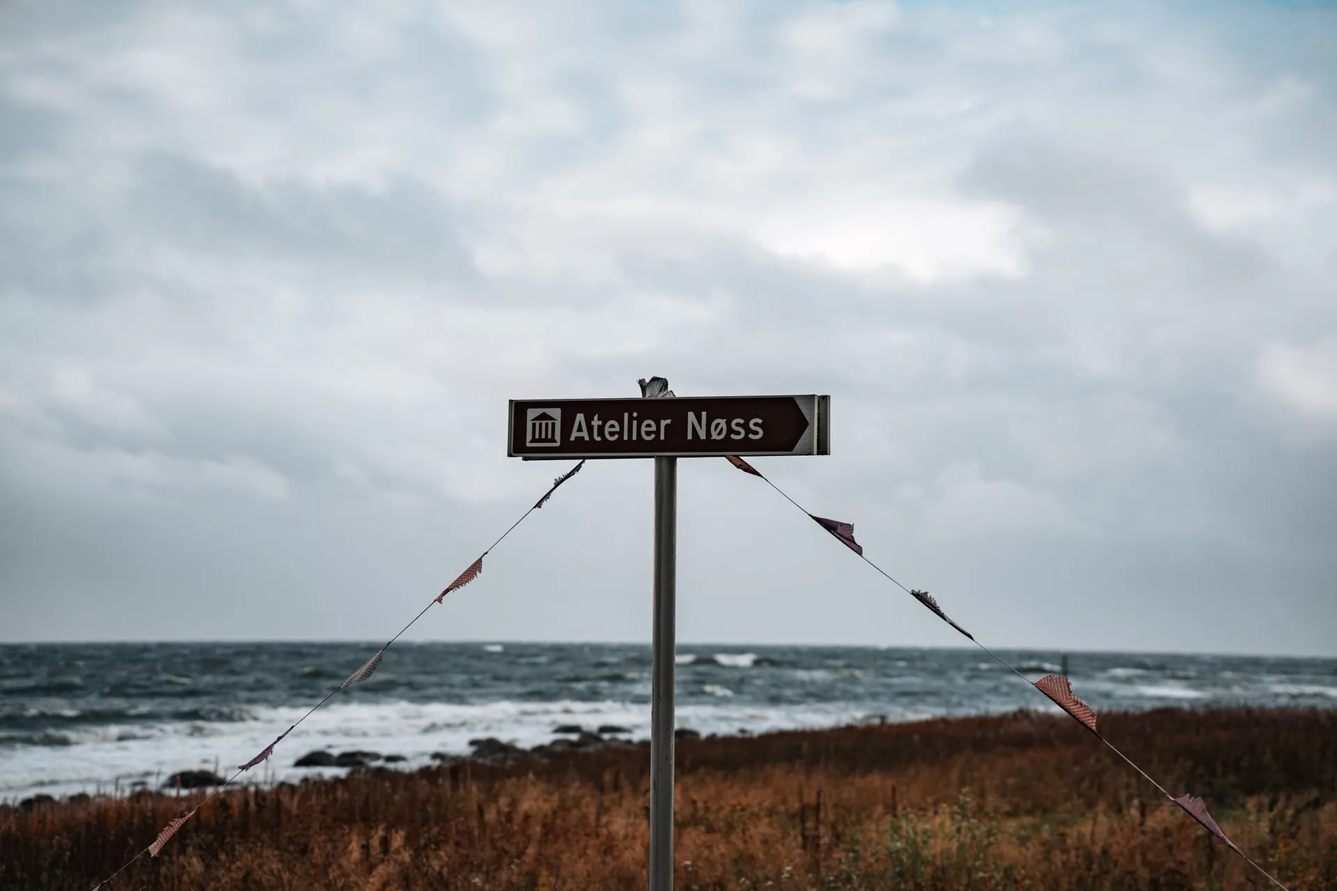 A brown signpost to Atelier Nøss with pennants on two strings, behind it the sea and brown grass