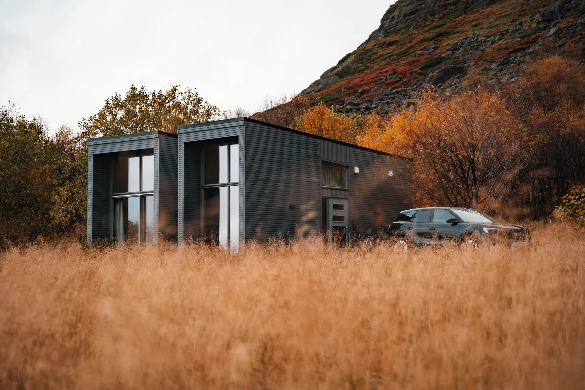 Our Airbnb in Nøss, a modern dark house with tall windows in golden grass, our Sixt rental car in front of it, orange trees and a rocky slope behind