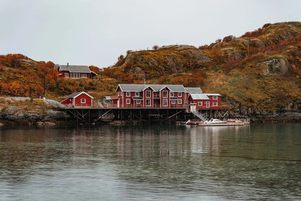 Rote Rorbuer auf Stelzen über dem ruhigen Wasser bei Sakrisøy, dahinter herbstlich orange Hügel
