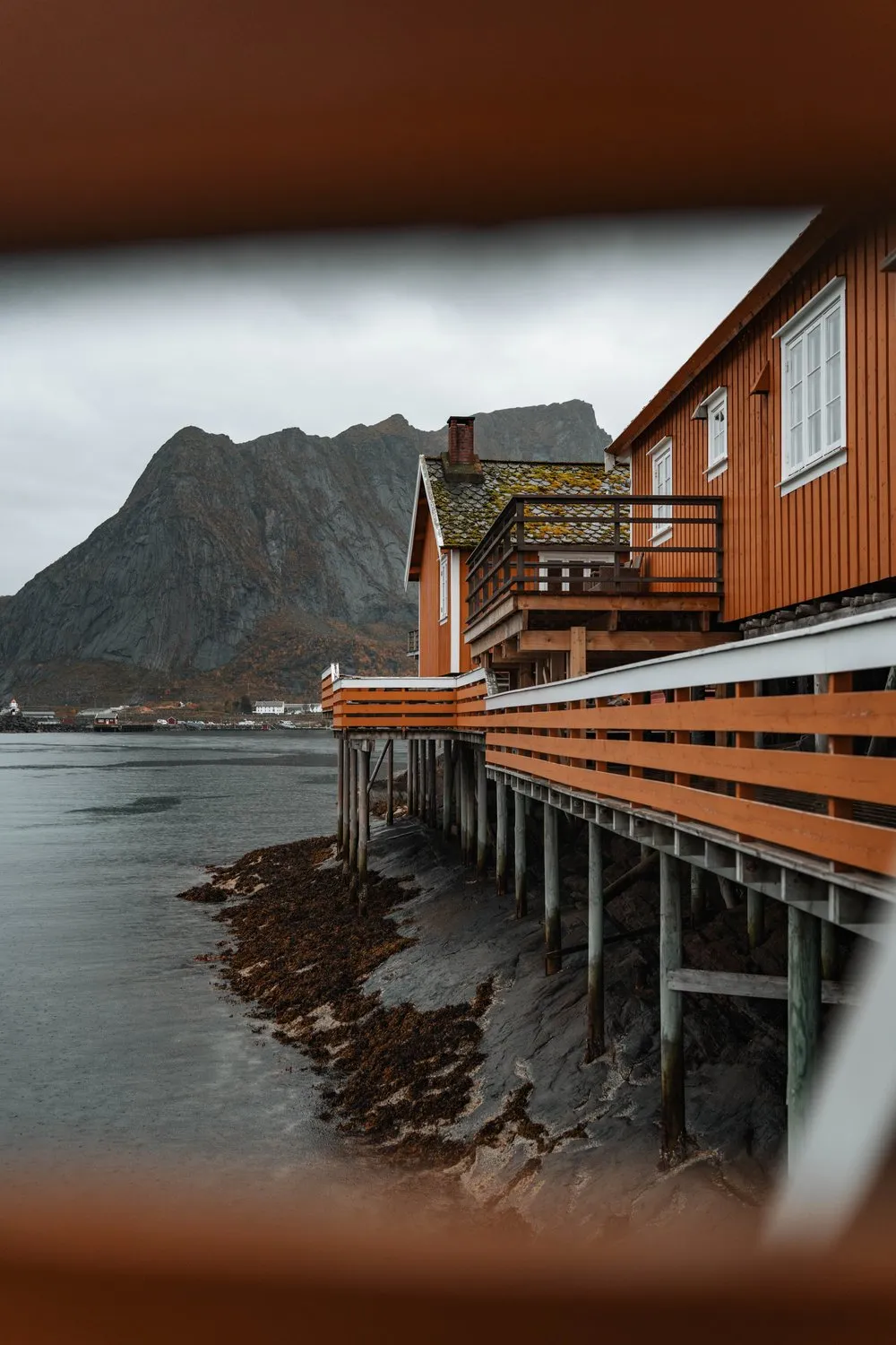 Through a gap between wooden beams onto the ochre rorbuer of Sakrisøy on their stilts by the water