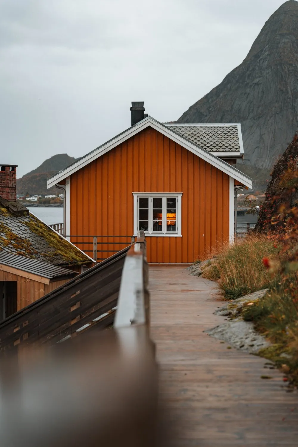 A wet wooden walkway leading to an ochre house in Sakrisøy, a pointed mountain behind