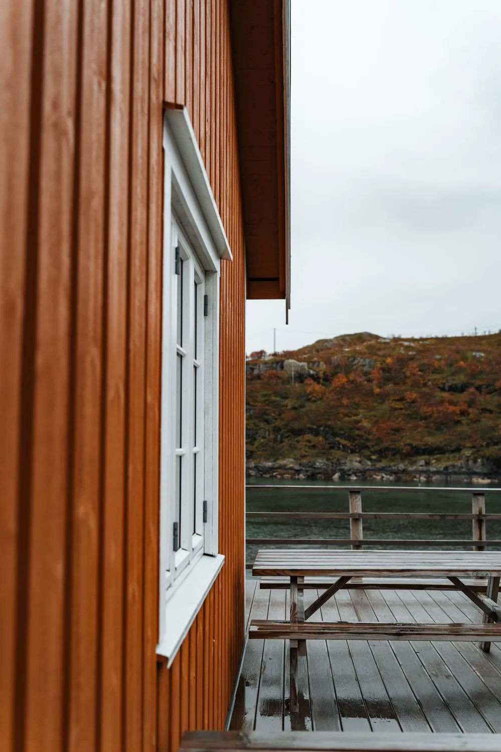 The ochre wooden wall of a rorbu in Sakrisøy with a white window frame, a wet picnic table on the deck beside it