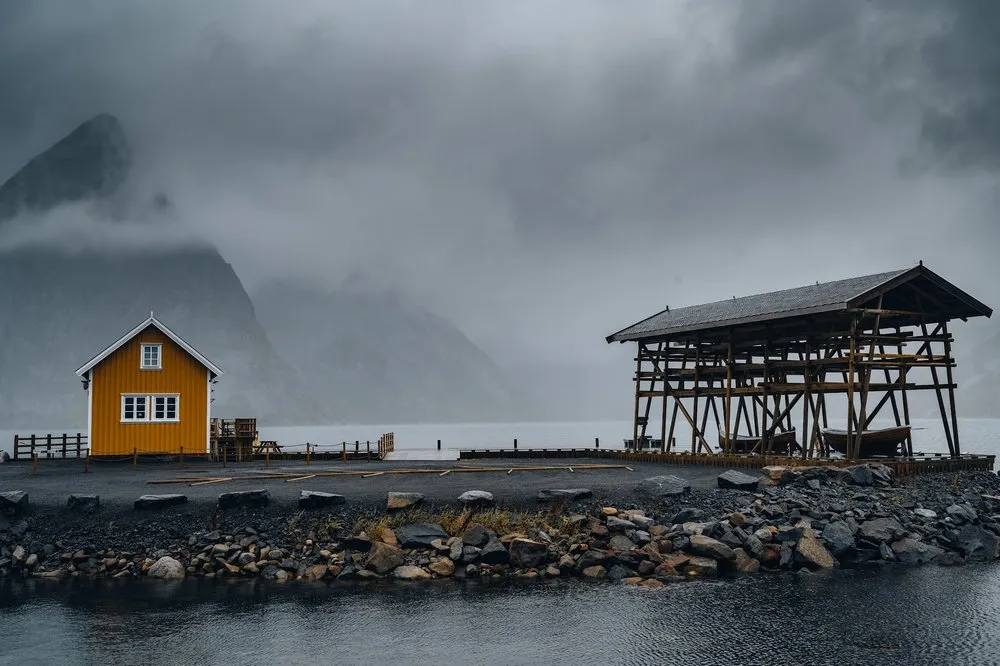 A yellow fishing house and a wooden frame with a roof at the water in Sakrisøy, a mountain in the fog behind