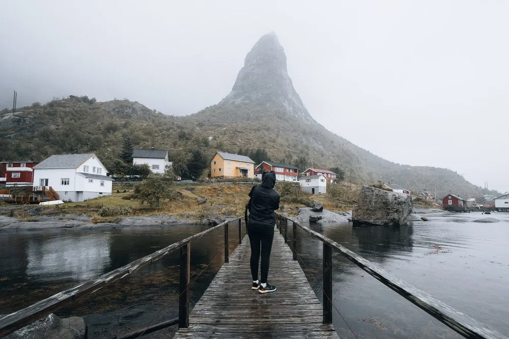 A person in a hood stands on a wooden footbridge in Reine and looks up at a pointed mountain above the houses