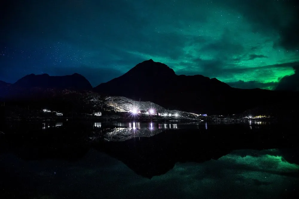 Grünes Nordlicht über einem dunklen Berg, der sich zusammen mit den Lichtern am Ufer im ruhigen Meer spiegelt