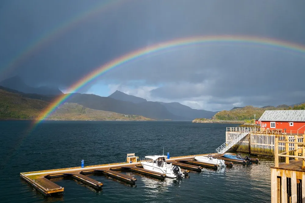 Der Regenbogen über dem Fjord, links ein Bootssteg mit Motorbooten, rechts eine rote Hütte