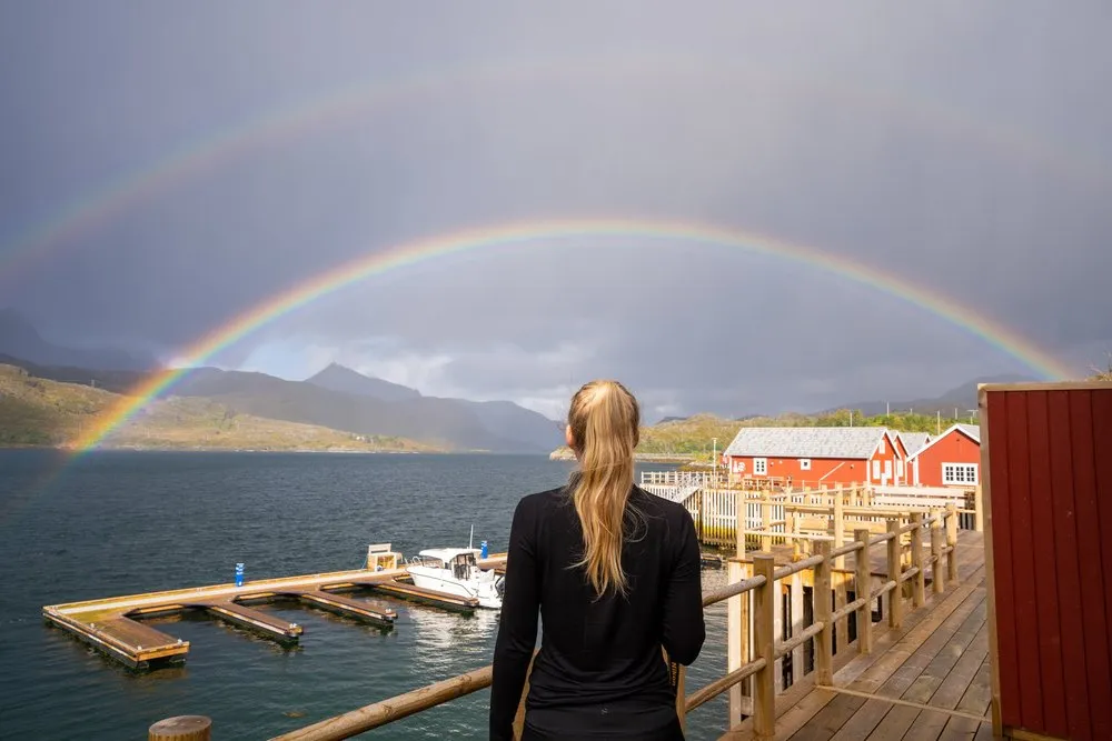 Eine Frau steht von hinten auf einem Holzsteg, vor ihr ein doppelter Regenbogen über Fjord und roten Hütten