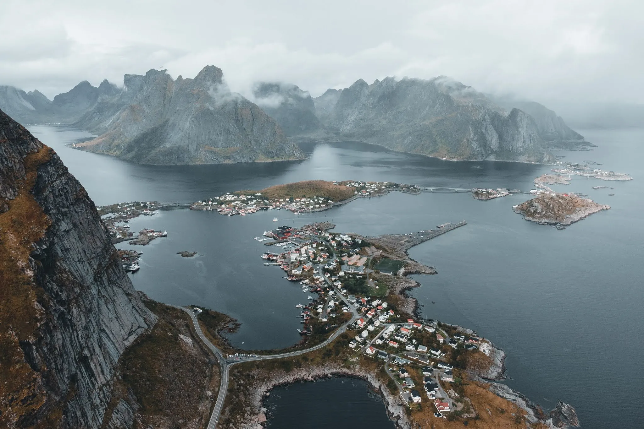 View from a great height onto a headland with small houses, a road and a fjord, mountains behind it in low clouds