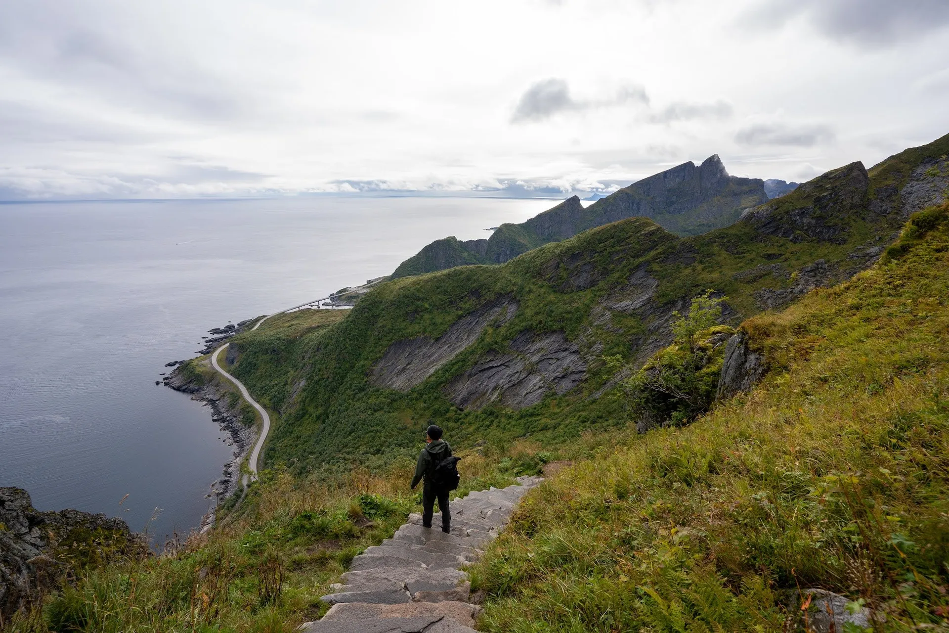 Eine Person mit Rucksack steht auf einer Steintreppe am grünen Hang, links das Meer und eine Strasse entlang der Küste
