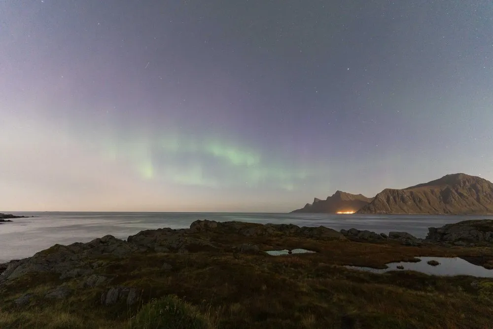 Schwache grüne Nordlichter über dem Meer und einem dunklen Berg, vorne Felsen und kleine Tümpel