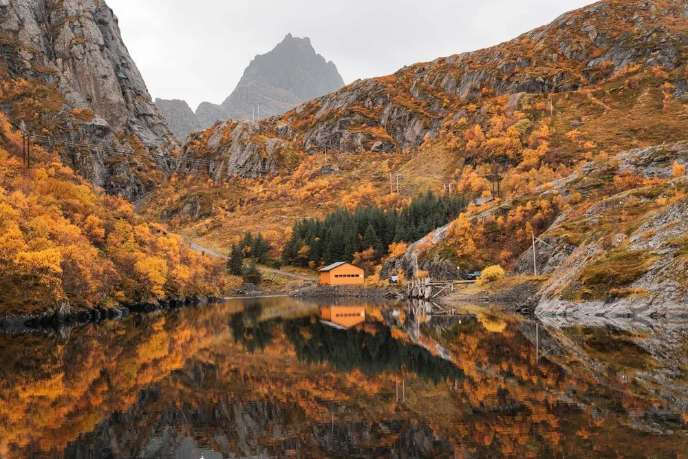 Ein oranges Haus am Ufer, im stillen Wasser spiegeln sich die herbstlich orangen Hänge und die grauen Gipfel