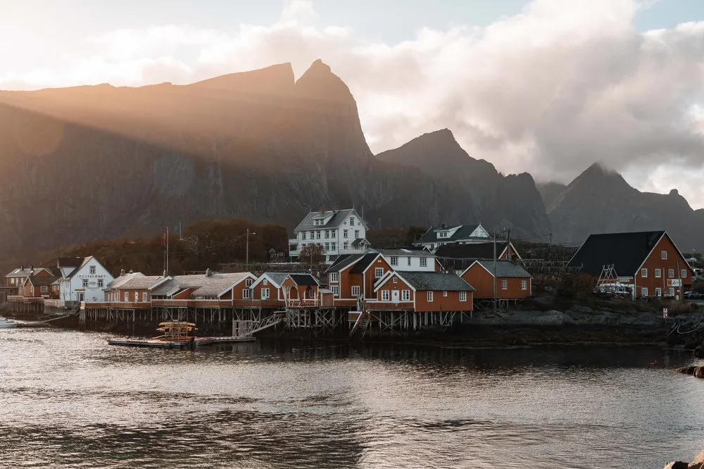 Die Rorbuer von Sakrisøy auf Stelzen am Wasser, dahinter Sonnenstrahlen über dem Berg