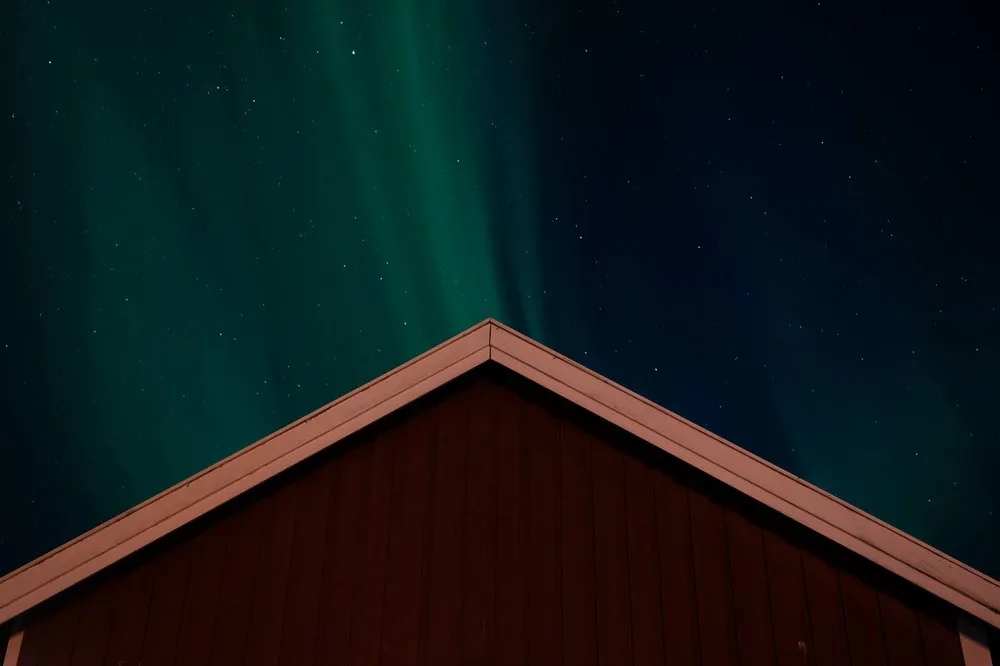 Green northern lights in a starry sky behind the gable of a red wooden cabin