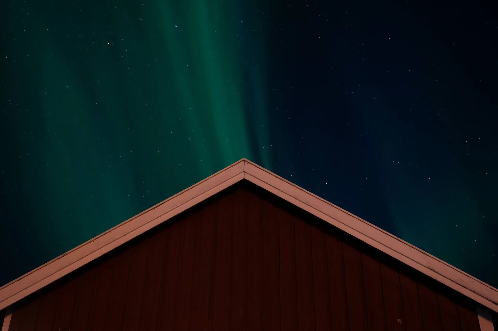 Grünes Nordlicht am Sternenhimmel hinter dem Giebel einer roten Holzhütte
