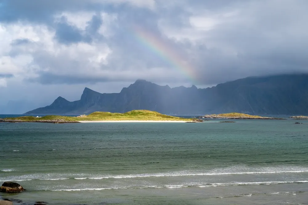 A rainbow above the mountains, turquoise water and a grass-covered island with a light strip of sand in front, near Fredvang