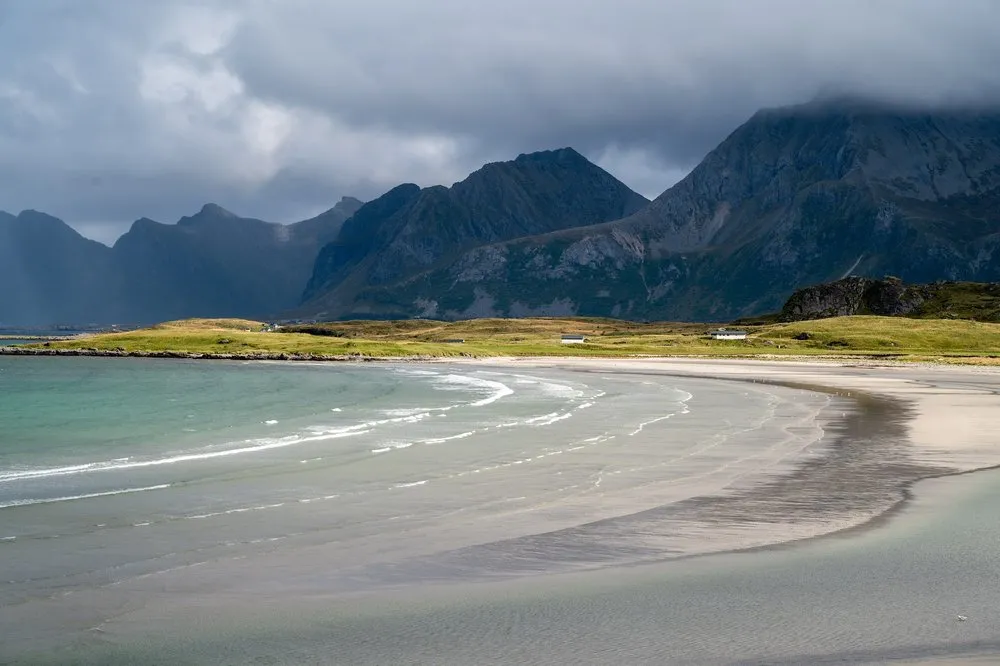 A curved light sandy beach with shallow water, dark mountains under clouds behind it