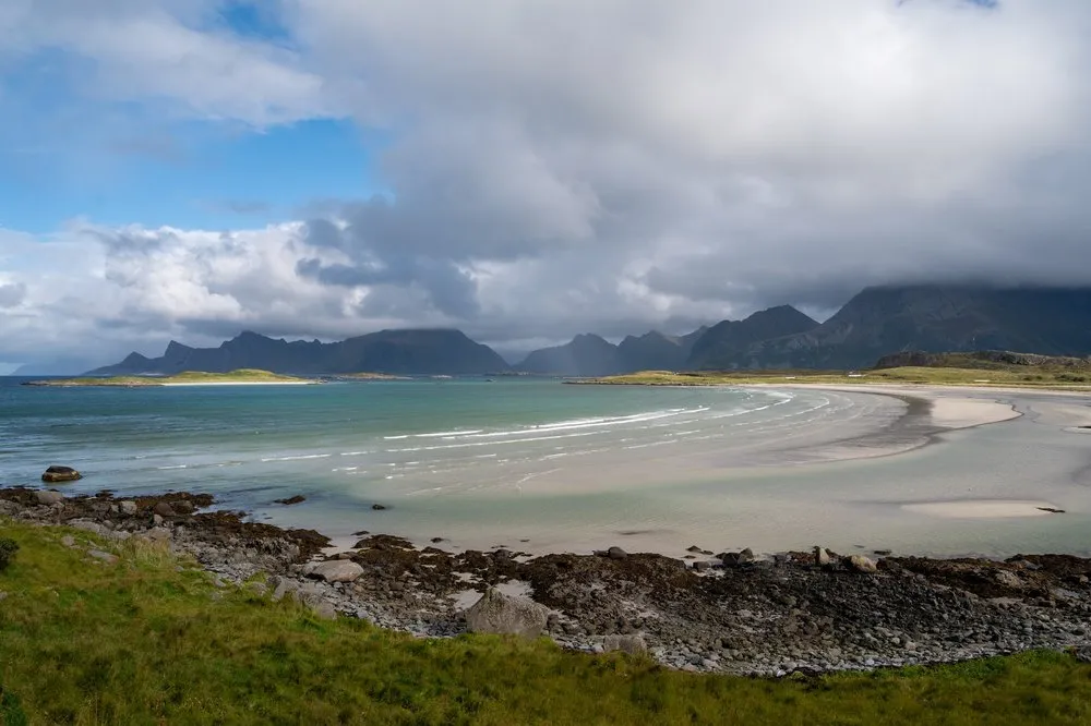A bay with light sand and turquoise water, stones and grass in the foreground, mountains and blue sky behind