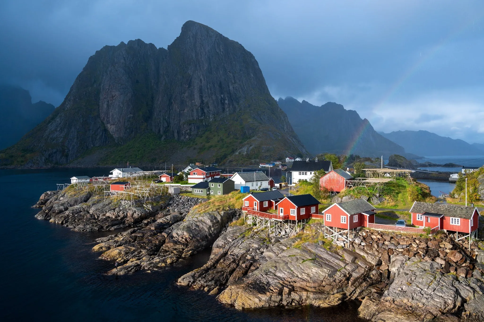 Red fishing cabins of Hamnøy on rocks by the water in front of a steep mountain, a rainbow behind them under a dark sky