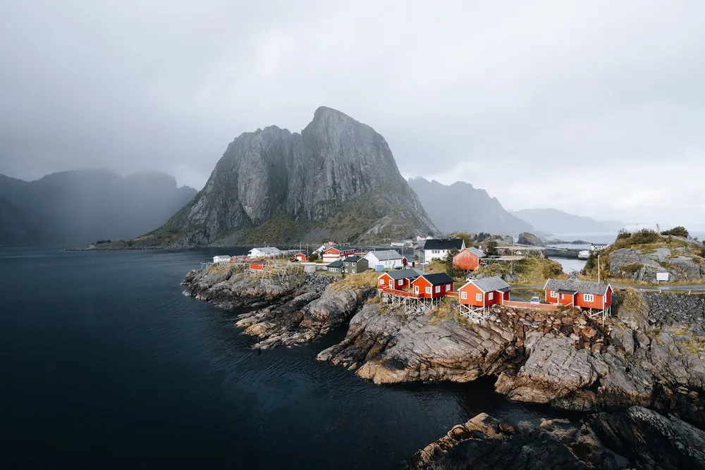 The red fishing cabins of Hamnøy on rocks in front of a steep mountain under a grey, hazy sky