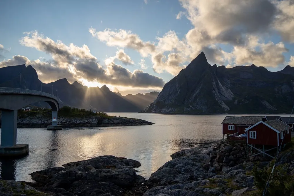 The sun hangs low between the mountains, a bridge on the left, red fishing cabins and a pointed mountain on the right at Hamnøy