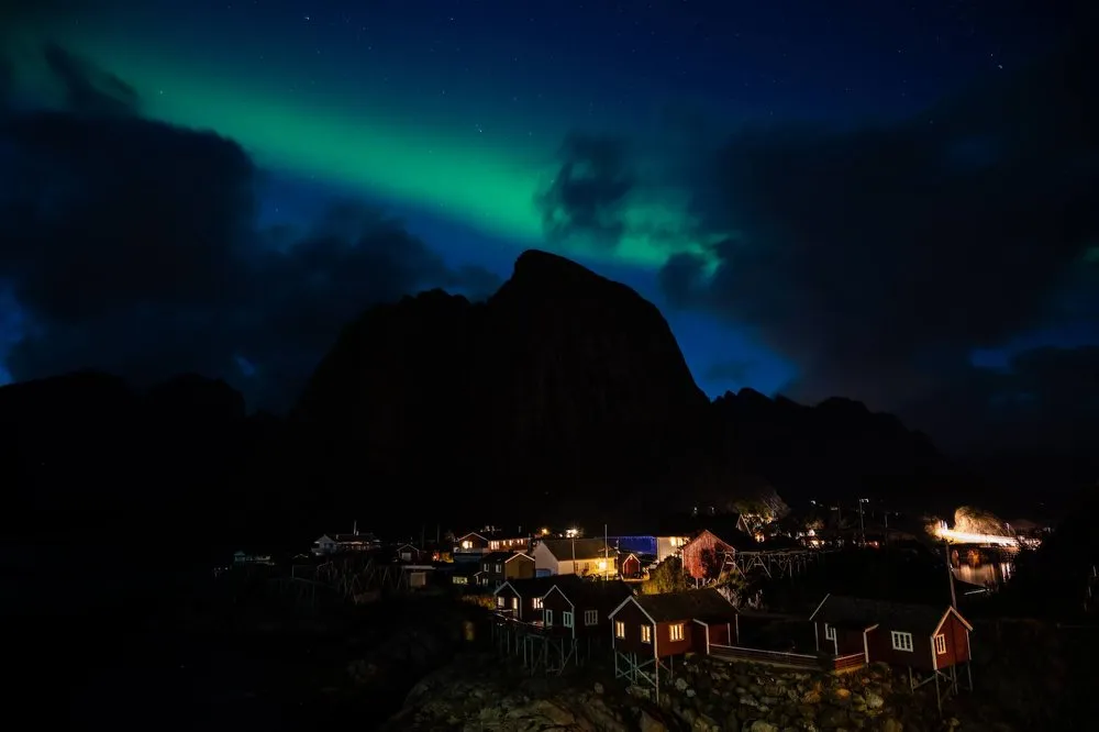 Green northern lights above the dark silhouette of the mountain, illuminated red little houses of Hamnøy below
