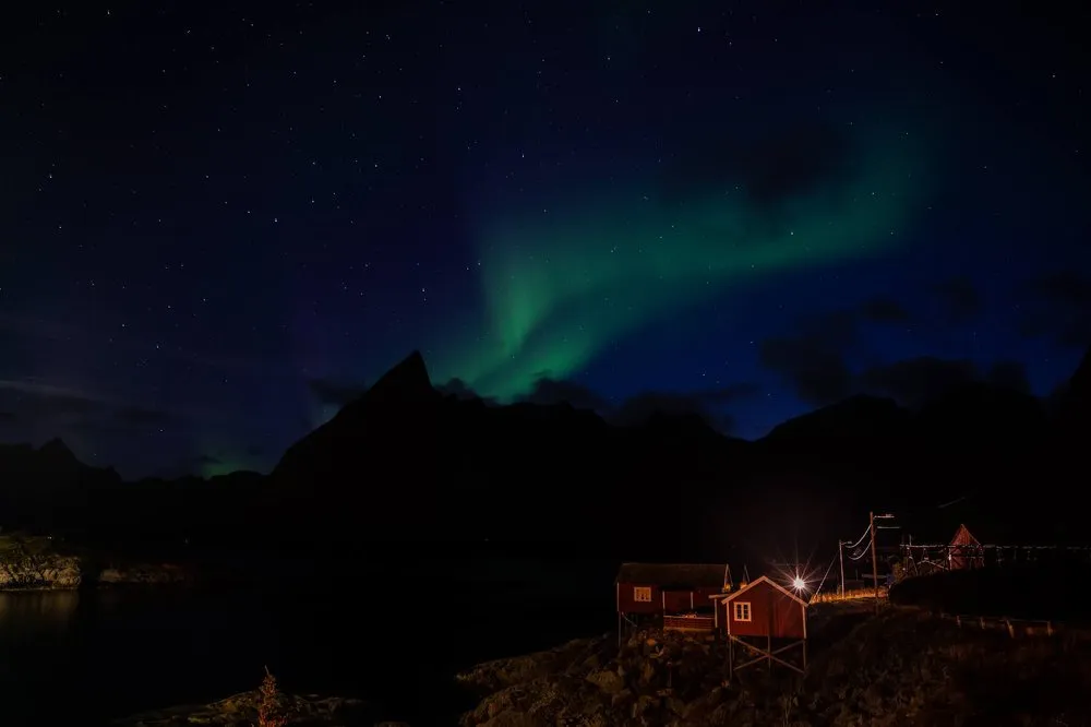 Northern lights behind a pointed mountain silhouette, two red cabins by the water below and a bright point of light