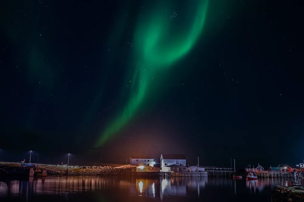 A green band of northern lights above the harbour, white buildings and street lamps in front, reflected in the water