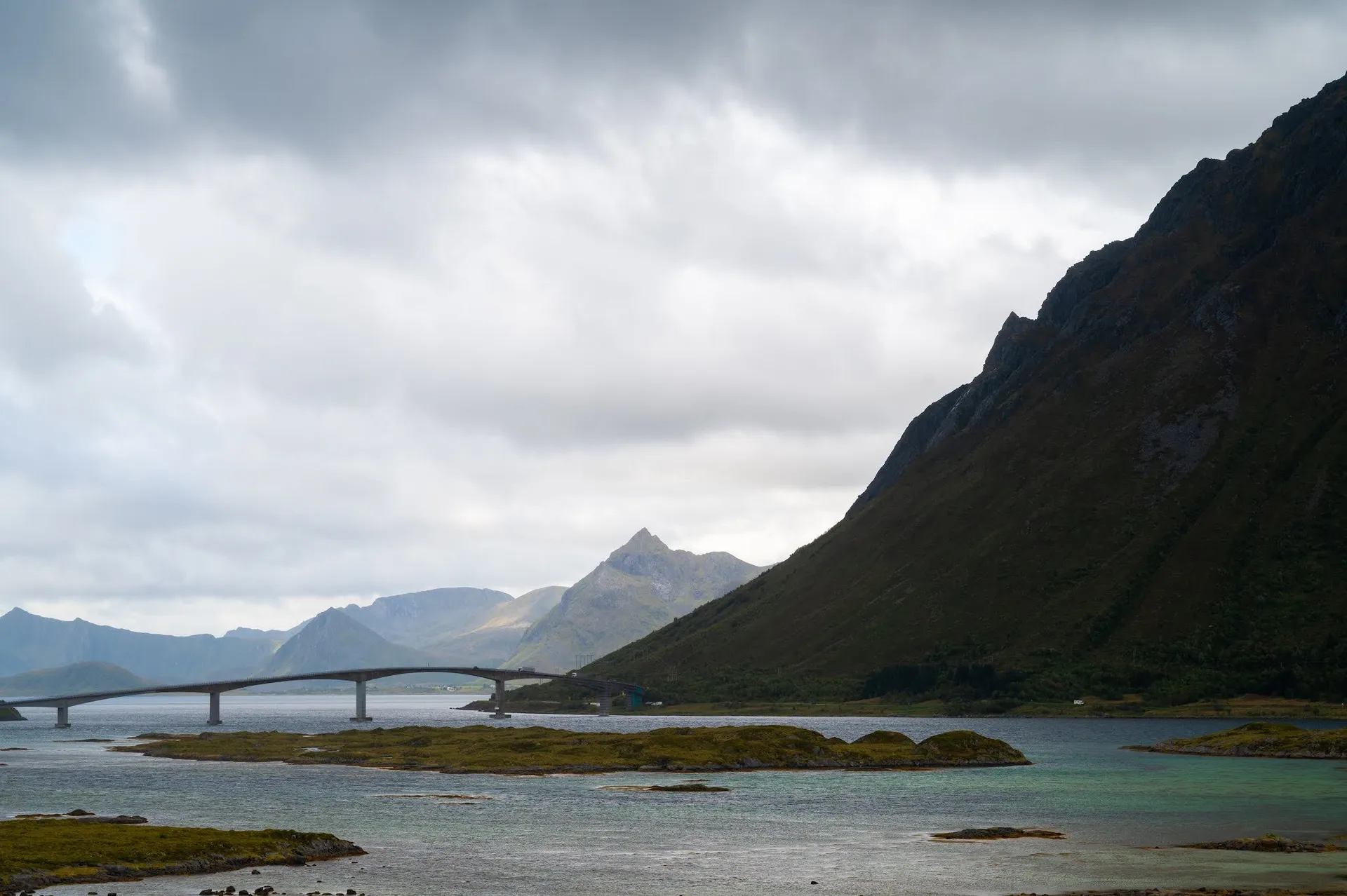 Eine lange, flach geschwungene Brücke über einen Meeresarm, rechts ein steil abfallender Berghang, links Berge im Dunst