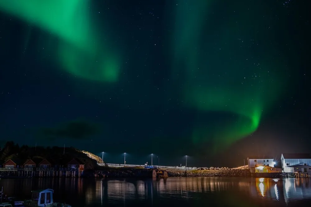 Green northern lights above the harbour, cabins under a hill on the left, white buildings on the right, everything reflected in the water