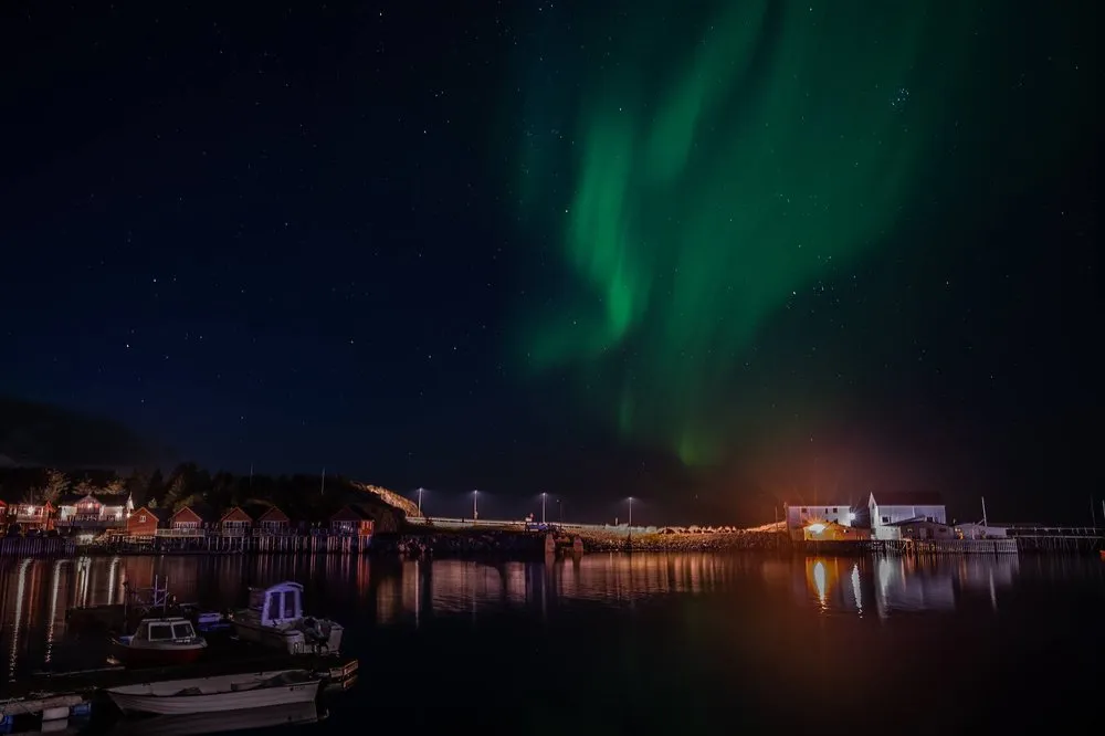 Northern lights on the right above the harbour, small boats in the foreground, stars in the dark sky