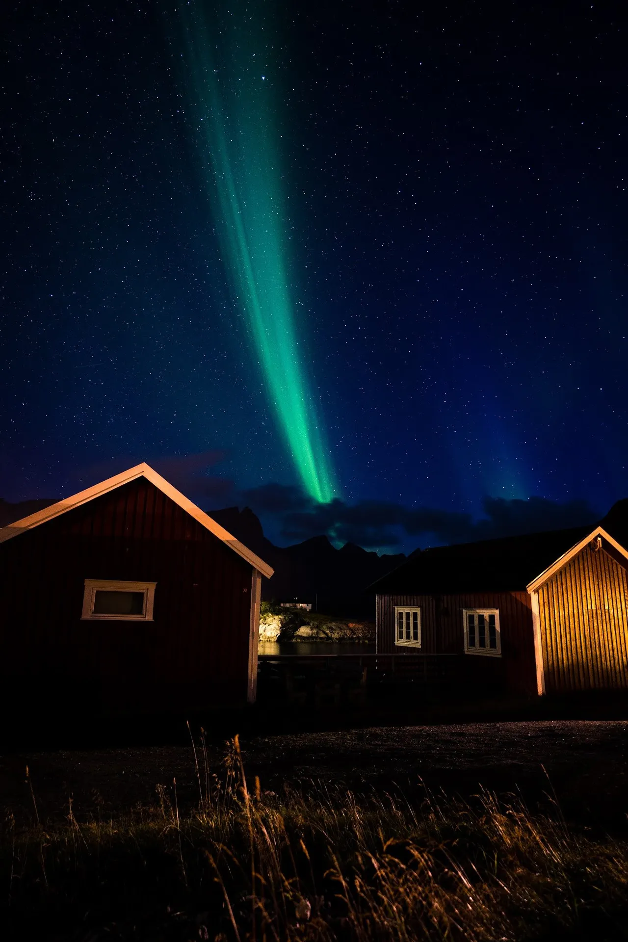 Ein schmaler grüner Lichtstrahl des Nordlichts steht zwischen zwei roten Hütten am Sternenhimmel