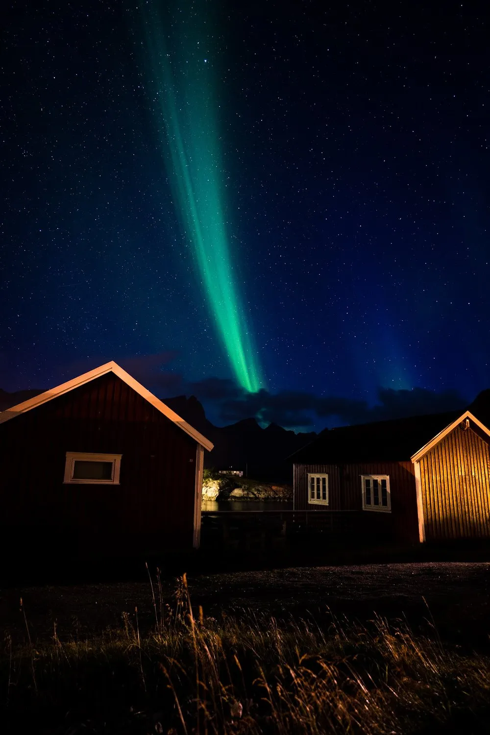 A narrow green beam of the northern lights stands between two red cabins in the starry sky