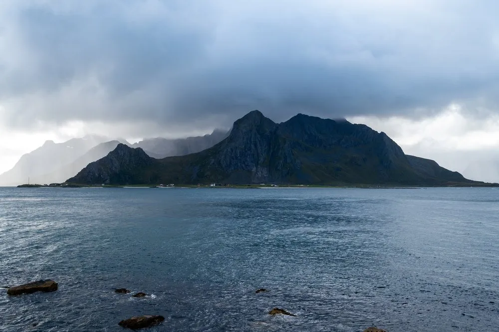 A dark mountain island in the sea under heavy rain clouds, a light strip of cloud in the background