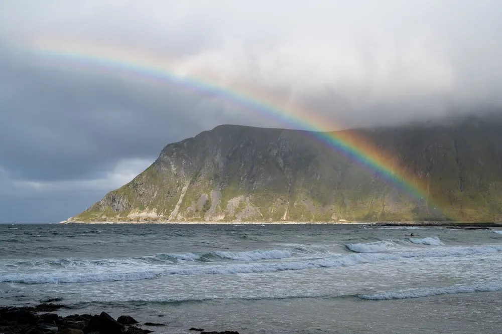A rainbow arches over a green mountainside at Skagsanden Beach, waves roll onto the beach in front