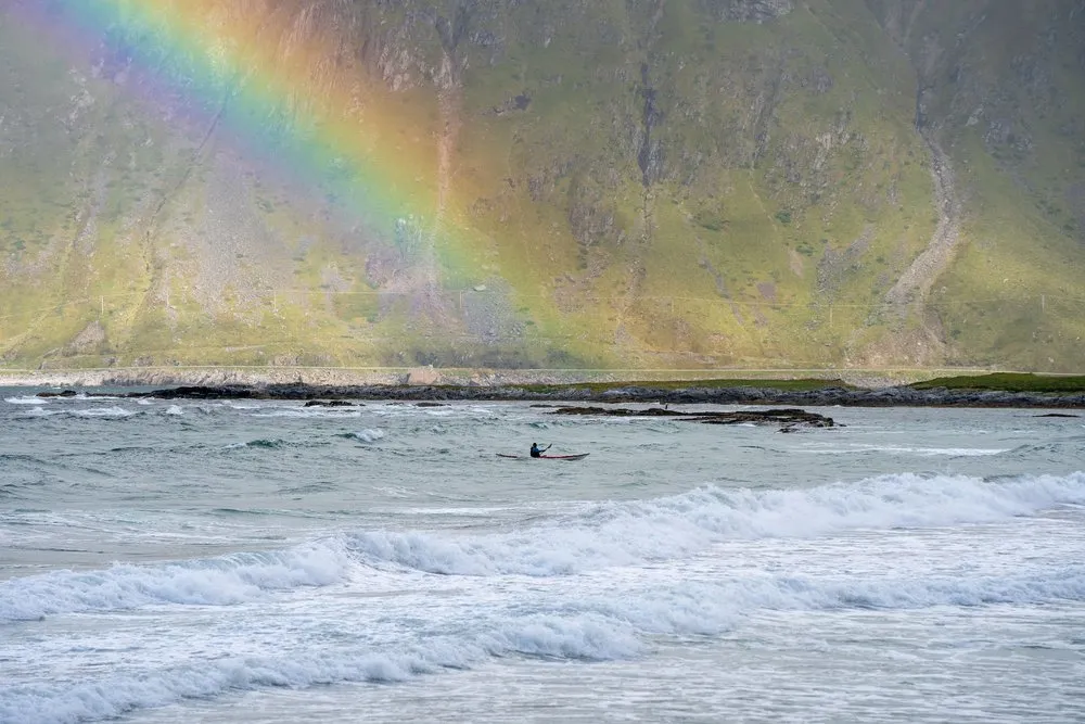 A kayaker in the surf at Skagsanden Beach, a mountainside with the end of a rainbow behind