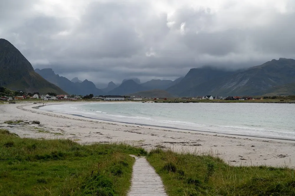 A wooden walkway leads through grass to a light sandy beach at Rambergstranda, houses and mountains in clouds behind