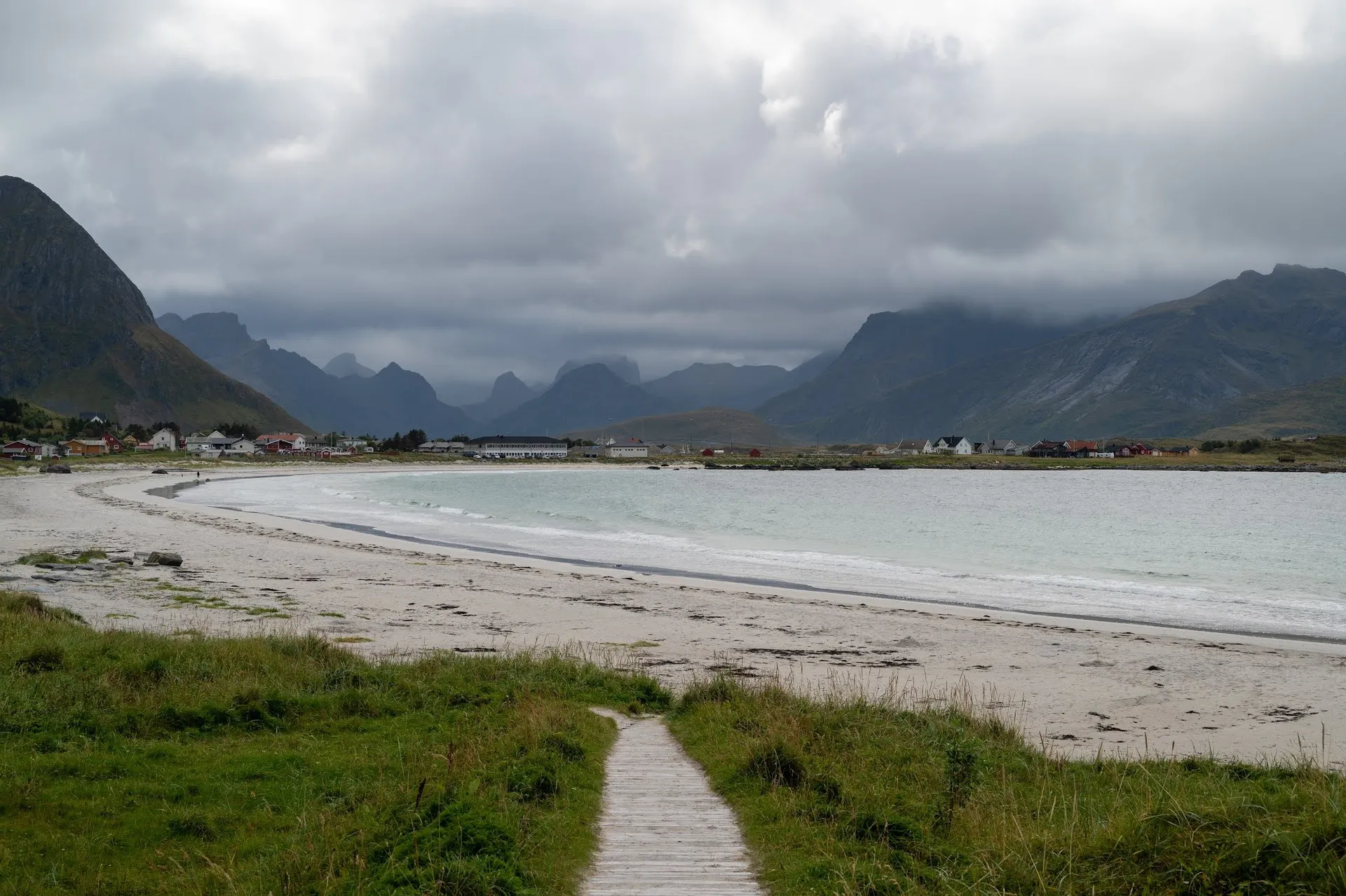 Ein Holzsteg führt durch Gras zu einem hellen Sandstrand am Rambergstranda, dahinter Häuser und Berge in Wolken