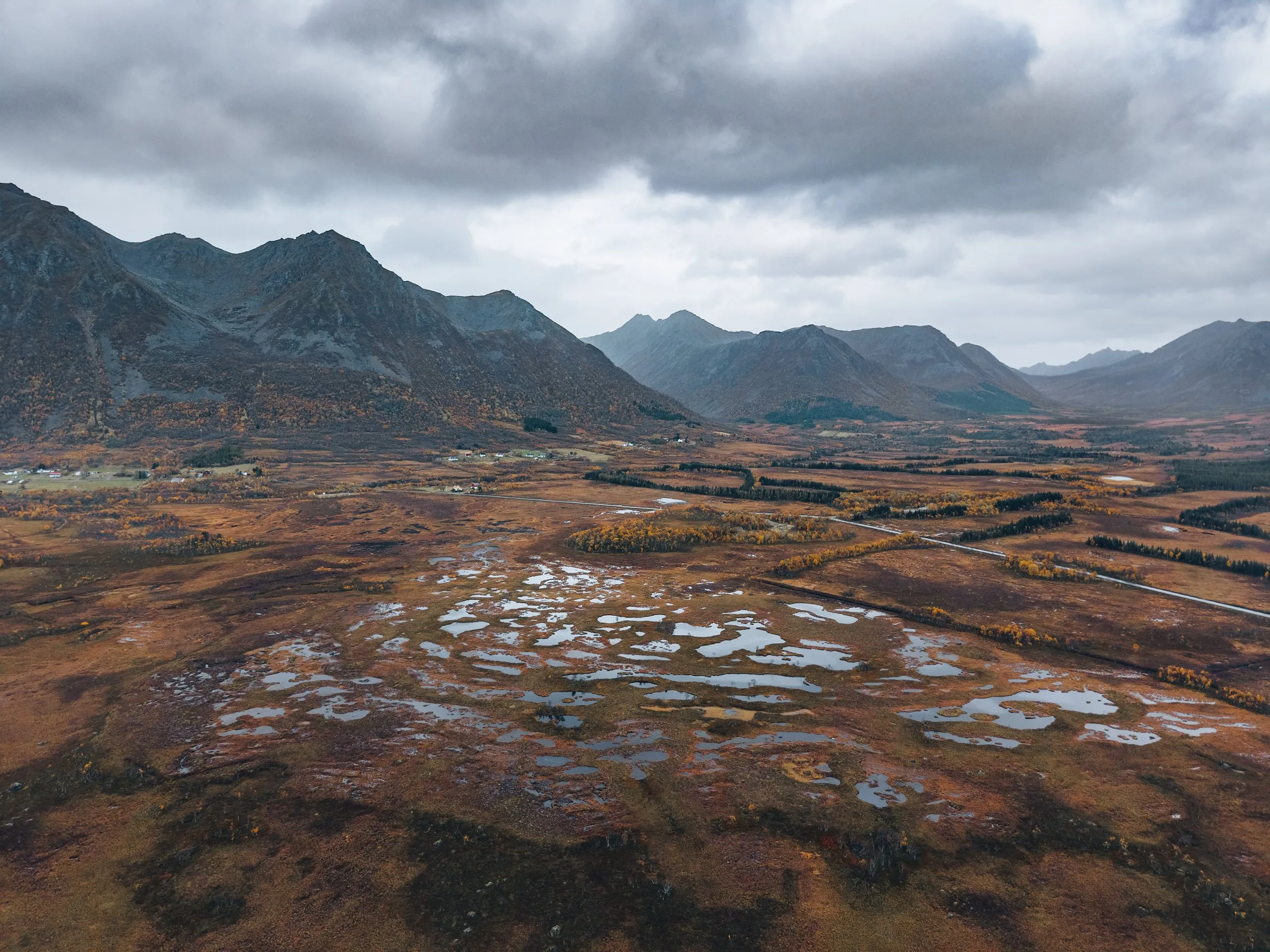 Reddish brown moorland with lots of small pools from the air, a mountain range under grey clouds behind, on the way to Andøya